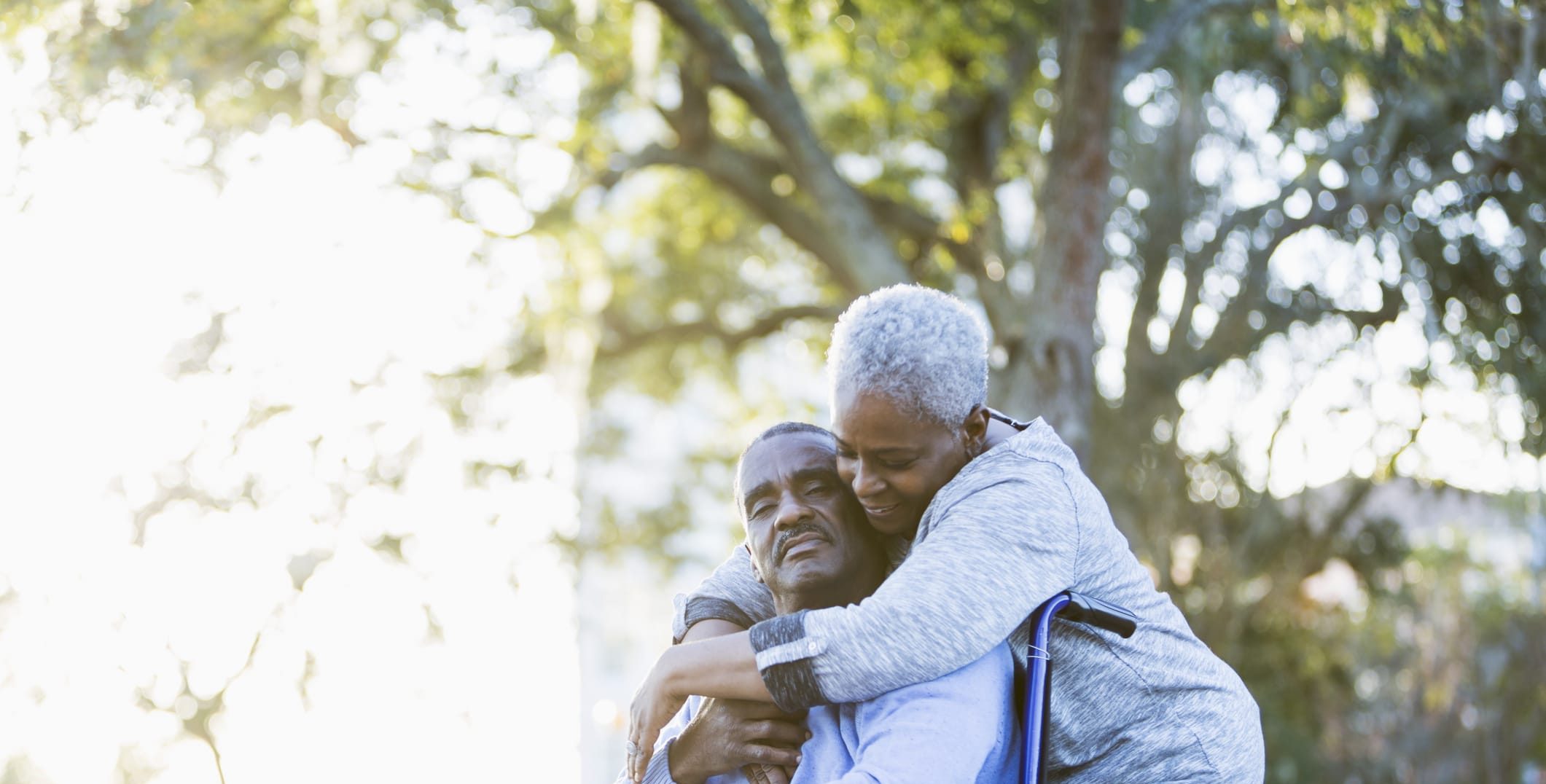 an older couple hugging outside on a sunny day, with one in a wheelchair